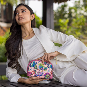A woman in a white outfit holding an Anuschka Fabric with Leather Trim Dome Cosmetic Bag - 13002 purse with a zip entry while looking thoughtfully off to the side.