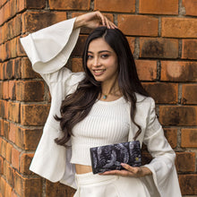 Load image into Gallery viewer, A smiling woman in a white outfit with zip pockets, holding a Fabric with Leather Trim Three-Fold RFID Wallet - 13007 by Anuschka against a brick wall background.
