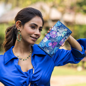 A woman in a blue blouse smiling while holding up a patterned, Anuschka Fabric with Leather Trim Three-Fold RFID Wallet - 13007.