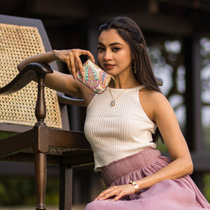 A woman in a sleeveless top and pleated skirt sitting by an outdoor table and posing with her hand on her cheek, near her Anuschka Fabric with Leather Trim Zip Travel Pouch - 13008.