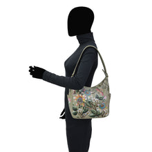 Load image into Gallery viewer, A person wearing a black outfit and carrying an Anuschka Classic Hobo With Side Pockets - 382 hand-painted artwork floral patterned genuine leather handbag stands in profile against a white background.

