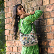 Load image into Gallery viewer, A woman in a green dress poses with a hand-painted, floral-patterned Anuschka Classic Hobo With Side Pockets - 382 handbag against a brick wall.
