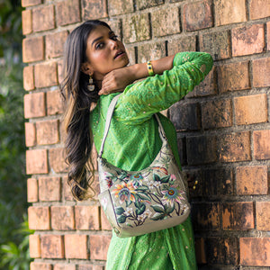 A woman in a green dress poses with a hand-painted, floral-patterned Anuschka Classic Hobo With Side Pockets - 382 handbag against a brick wall.