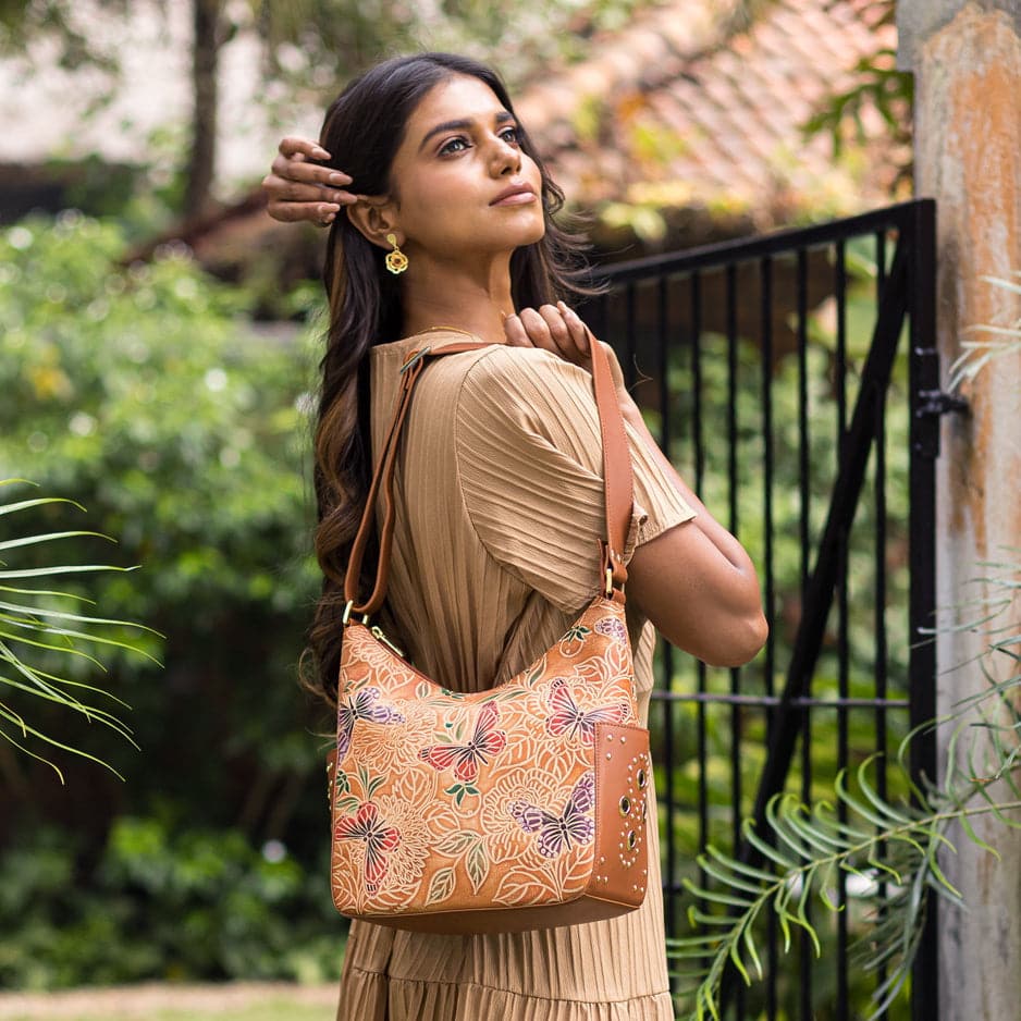 A woman poses with an Anuschka Classic Hobo With Studded Side Pockets - 433, adjusting her hair and looking away contemplatively.