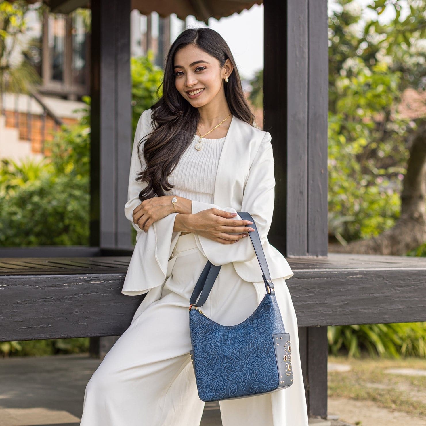 A woman in a white outfit holding an Anuschka Classic Hobo With Studded Side Pockets - 433 handbag sits on a bench with foliage in the background.