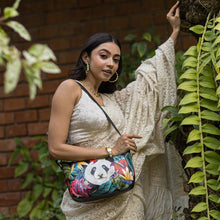 Load image into Gallery viewer, A woman in a white outfit holding an Anuschka Everyday Shoulder Hobo -670 with a panda design poses beside green foliage.
