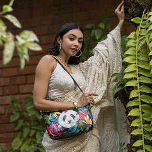 A woman in a white outfit holding an Anuschka Everyday Shoulder Hobo -670 with a panda design poses beside green foliage.