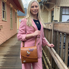 Load image into Gallery viewer, Woman in pink suit posing with an Anuschka Flap Messenger Crossbody - 692, standing on a wooden deck.
