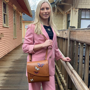 Woman in pink suit posing with an Anuschka Flap Messenger Crossbody - 692, standing on a wooden deck.