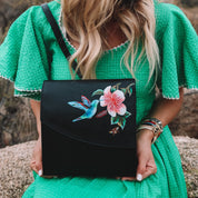 Woman in a green dress holding an Anuschka Flap Messenger Crossbody - 692 with a hummingbird design.