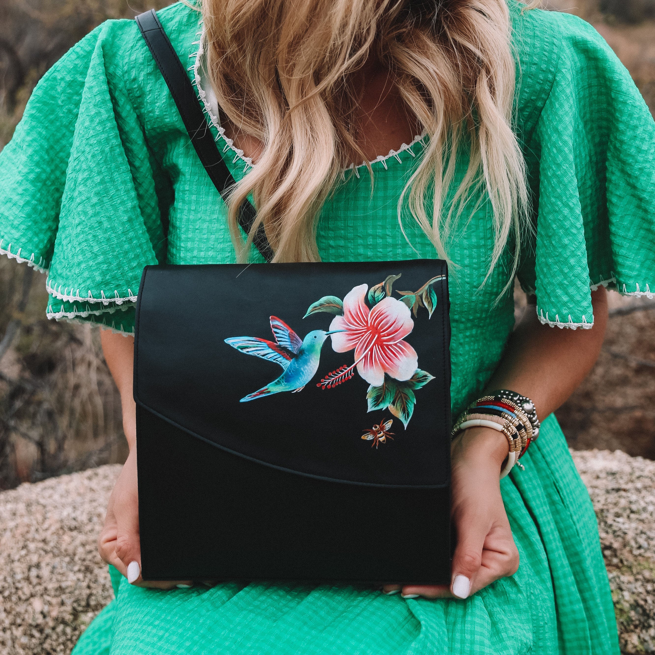 Woman in a green dress holding an Anuschka Flap Messenger Crossbody - 692 with a hummingbird design.