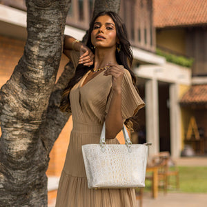 A woman in an elegant beige dress posing with an Anuschka Medium Tote - 693 featuring a zippered pocket next to a tree.