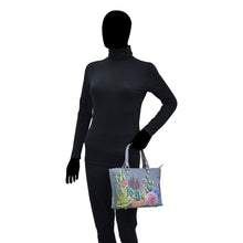 Load image into Gallery viewer, Mannequin in a black turtleneck and gloves holding a colorful floral, hand painted Anuschka Medium Tote - 693 with a zippered compartment.
