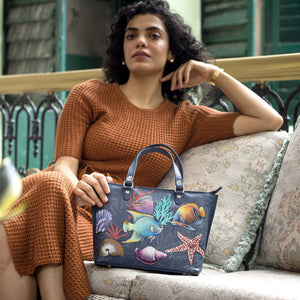 A woman sitting leisurely on a cushioned bench holding an Anuschka genuine leather Medium Tote - 693 with a hand-painted, colorful fish design.