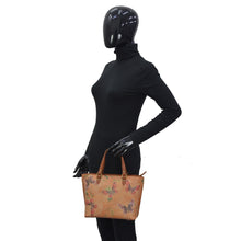 Load image into Gallery viewer, Mannequin dressed in a black turtleneck and gloves, wearing a motorcycle helmet, holding an Anuschka Medium Tote - 693 with a floral pattern.
