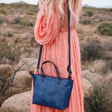 Load image into Gallery viewer, A person in a coral dress holding an Anuschka blue patterned leather tote (Medium Tote - 693) stands in a desert landscape.
