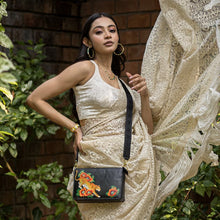 Load image into Gallery viewer, A woman posing elegantly in a cream dress accessorized with earrings and a necklace, holding an Anuschka Triple Compartment Crossbody - 696 with colorful embroidery.
