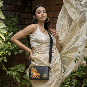 A woman posing elegantly in a cream dress accessorized with earrings and a necklace, holding an Anuschka Triple Compartment Crossbody - 696 with colorful embroidery.