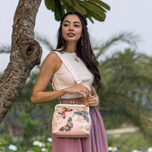 Load image into Gallery viewer, A woman standing outdoors, holding a pastel Anuschka Medium Frame Crossbody - 700 handbag with butterfly designs, wearing a white top and pleated skirt.
