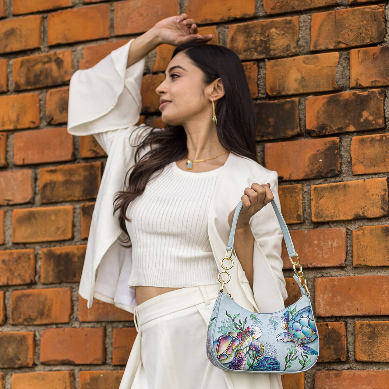 A woman in a white outfit poses with an Anuschka Small Convertible Hobo - 701 against a brick wall.