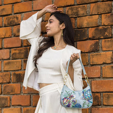 Load image into Gallery viewer, A woman in a white outfit poses with an Anuschka Small Convertible Hobo - 701 against a brick wall.
