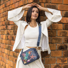 Load image into Gallery viewer, A woman in a white outfit posing with an Anuschka Twin Top Messenger - 704 purse with an adjustable strap against a brick wall.

