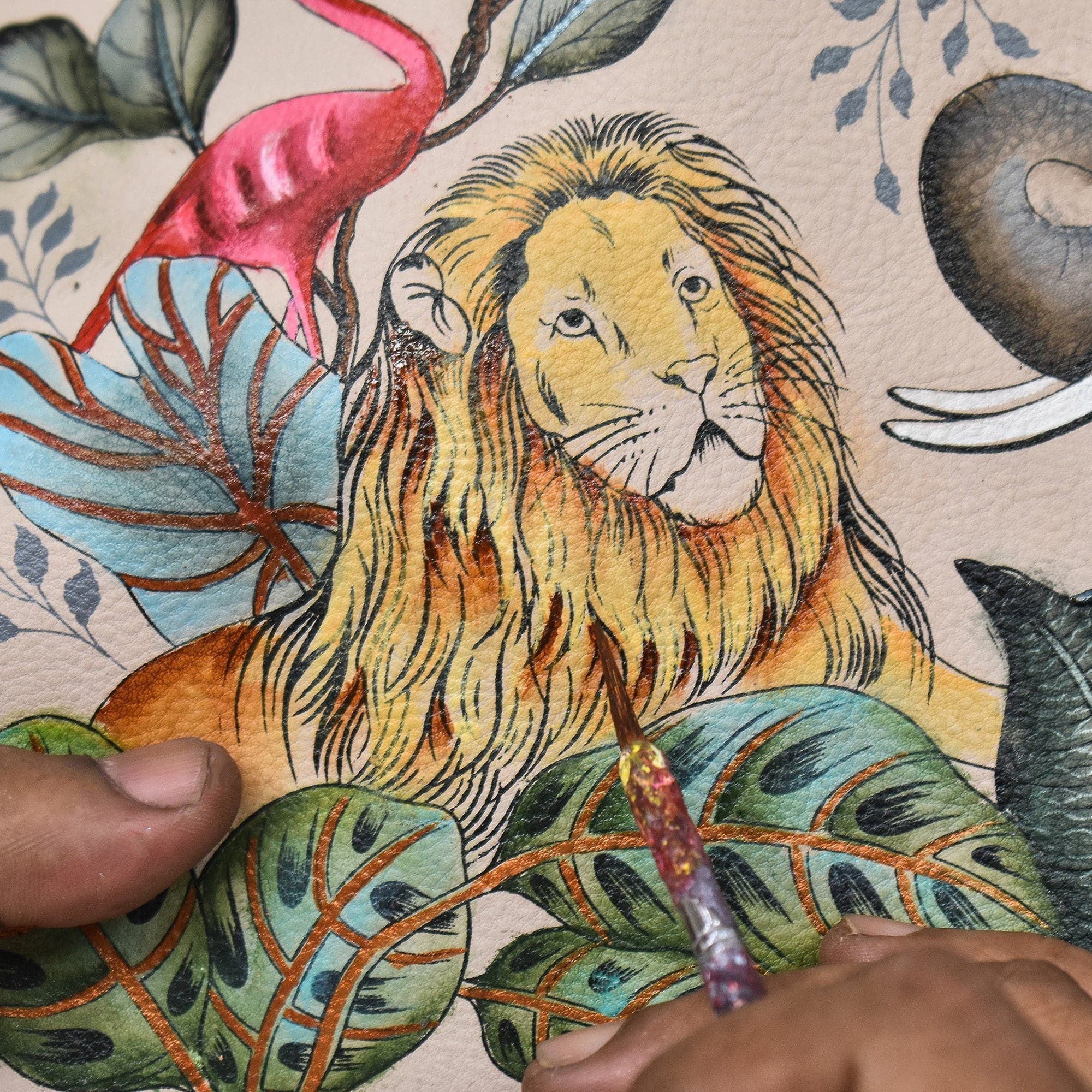 An Anuschka artist's hand painting a detailed lion surrounded by colorful flora on vintage-inspired paper.