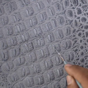 A person engraving a pattern onto the gray surface of a genuine leather Anuschka Organizer Wallet Crossbody - 1149 with a carving tool.