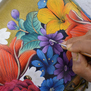A person is meticulously hand painting a colorful floral design on a genuine Anuschka Double Eyeglass Case - 1009 using a fine brush.