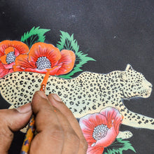Load image into Gallery viewer, A hand holding a paintbrush applies color to an illustration of a leopard intertwined with red poppies on an Anuschka Triple Compartment Crossbody - 696.

