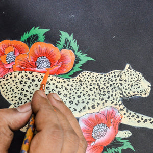 A hand holding a paintbrush applies color to an illustration of a leopard intertwined with red poppies on an Anuschka Triple Compartment Crossbody - 696.