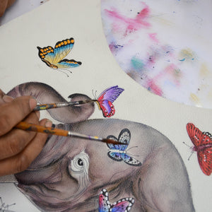 An artist's hand painting colorful butterflies on an Anuschka Slim Crossbody With Front Zip - 452 with an elephant illustration.