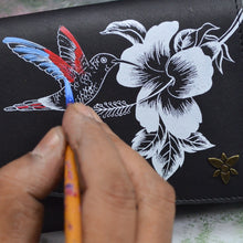Load image into Gallery viewer, An artist&#39;s hand painting a colorful hummingbird on an Anuschka Accordion Flap Wallet - 1174 with a black and white floral patterned surface, featuring RFID protection.
