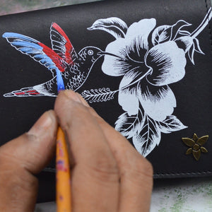 An artist's hand painting a colorful hummingbird on an Anuschka Accordion Flap Wallet - 1174 with a black and white floral patterned surface, featuring RFID protection.