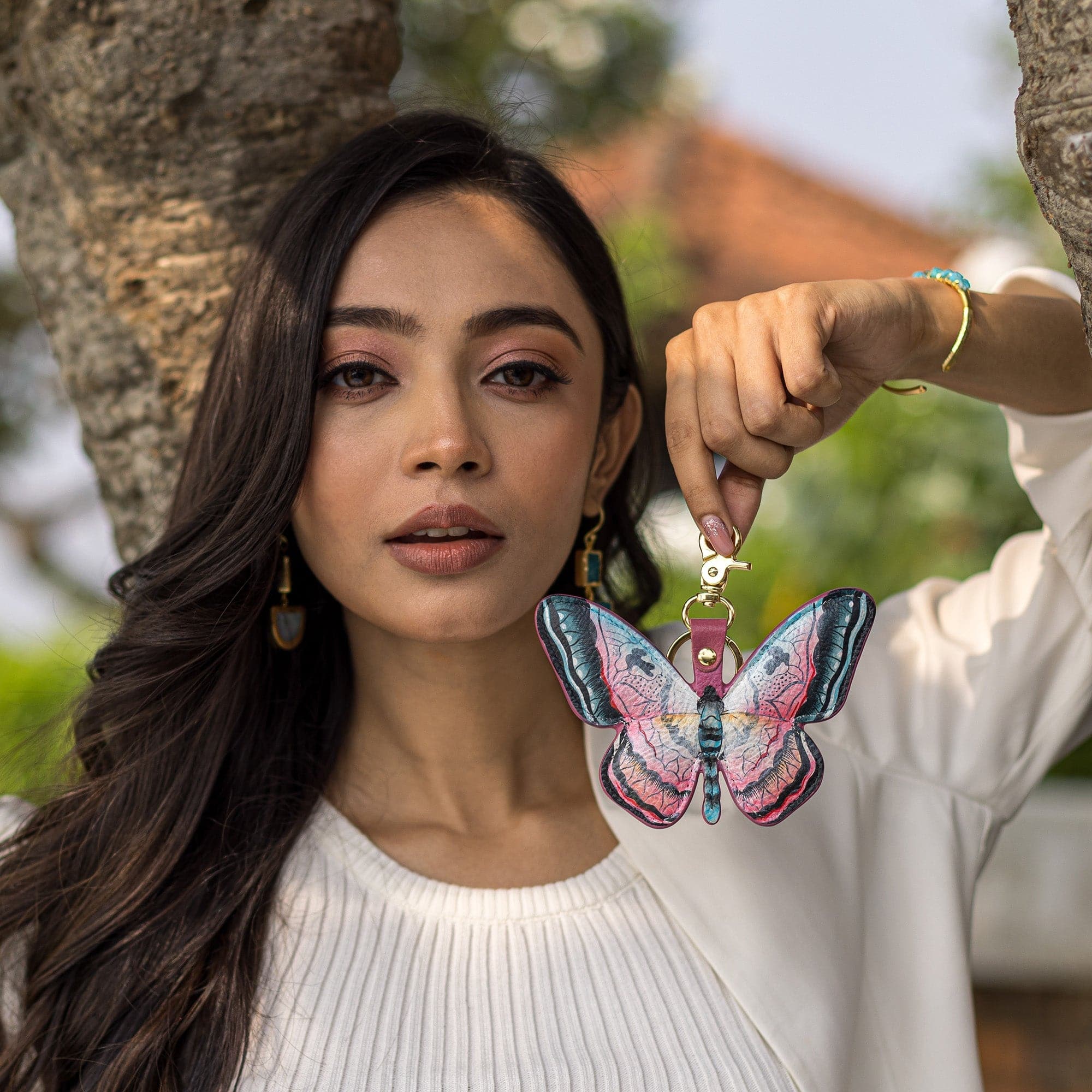 A woman holding a Painted Leather Bag Charm K0005 - Keycharms in the shape of a butterfly in front of a tree, by Anuschka.