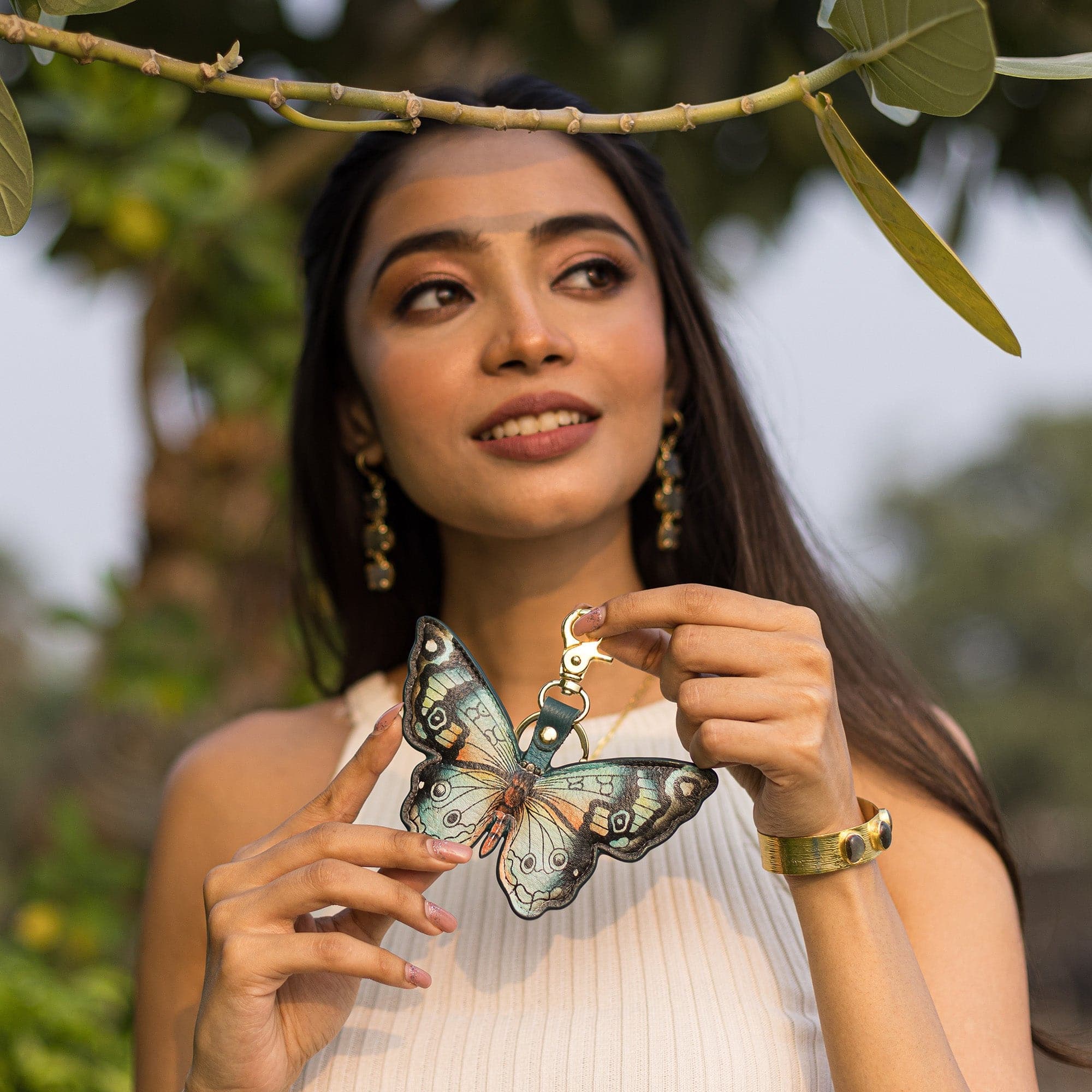 A woman holding a Painted Leather Bag Charm K0007 - Keycharms in the shape of a butterfly under a tree branch. Brand: Anuschka.