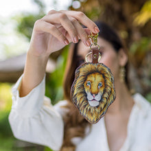 Load image into Gallery viewer, A person holding an Anuschka lion-faced, hand-painted keychain made of genuine leather in front of a blurred natural background.
