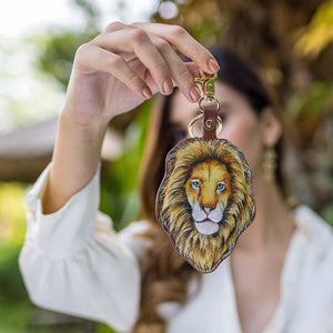 A person holding an Anuschka lion-faced, hand-painted keychain made of genuine leather in front of a blurred natural background.