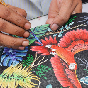 Close-up of hands painting a colorful parrot on a Anuschka Small Convertible Hobo - 701.