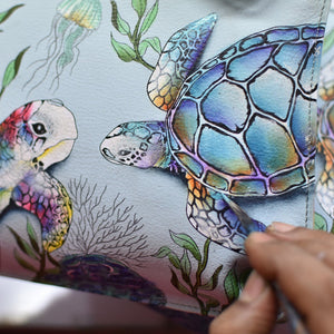 An artist's hand adding color to a sea turtle illustration on an Anuschka Triple Compartment Crossbody Organizer - 412 made of genuine leather.