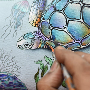 Paintbrush adding color to an illustrated sea turtle on an Anuschka Medium Zip Pouch - 1107 in a wildlife-themed artwork.