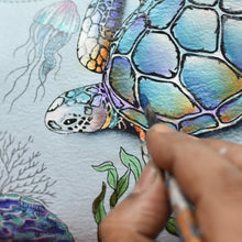 Load image into Gallery viewer, A close-up of a hand painting a colorful sea turtle on Anuschka hand painted leather, with intricate details and jellyfish in the background.
